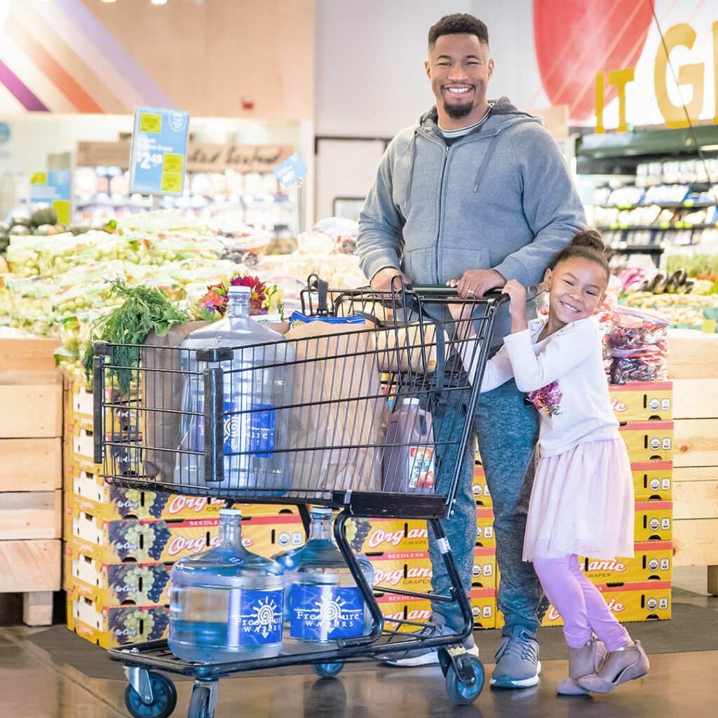 A father and his daughter are smiling at the camera as they shop for groceries and refill on bulk water. 